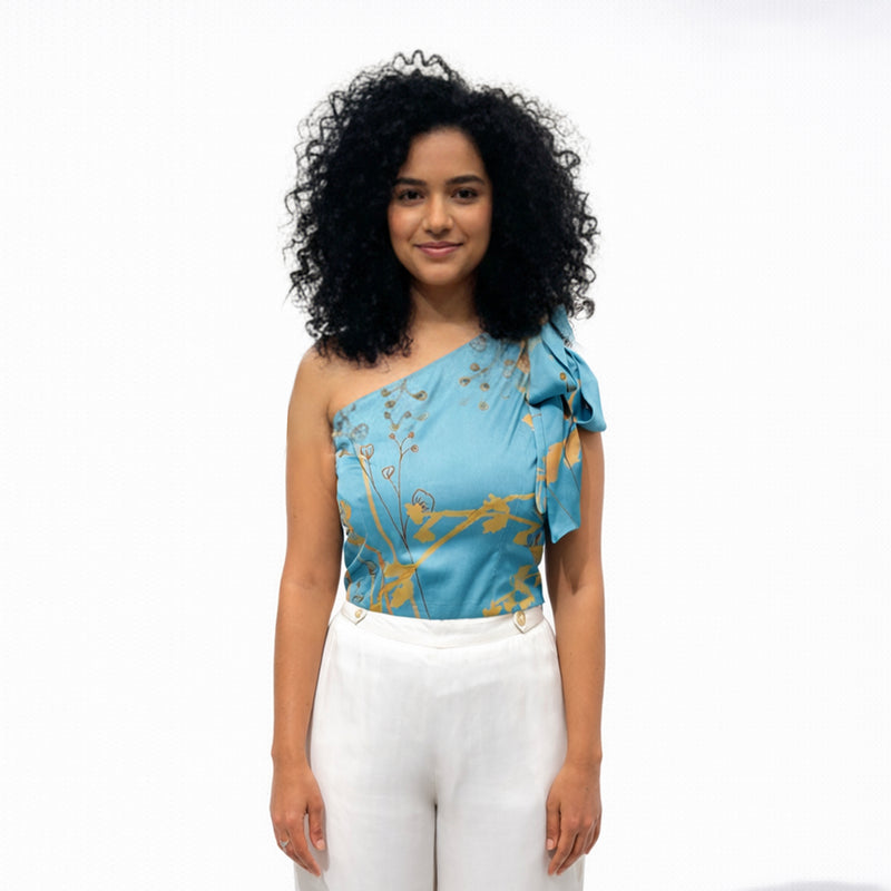 Front view of the teal one-shoulder floral print top styled with white wide-leg trousers