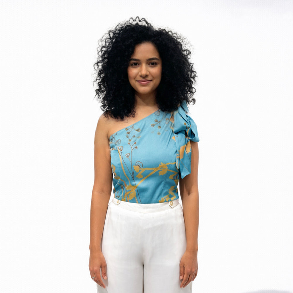 Front view of the teal one-shoulder floral print top styled with white wide-leg trousers
