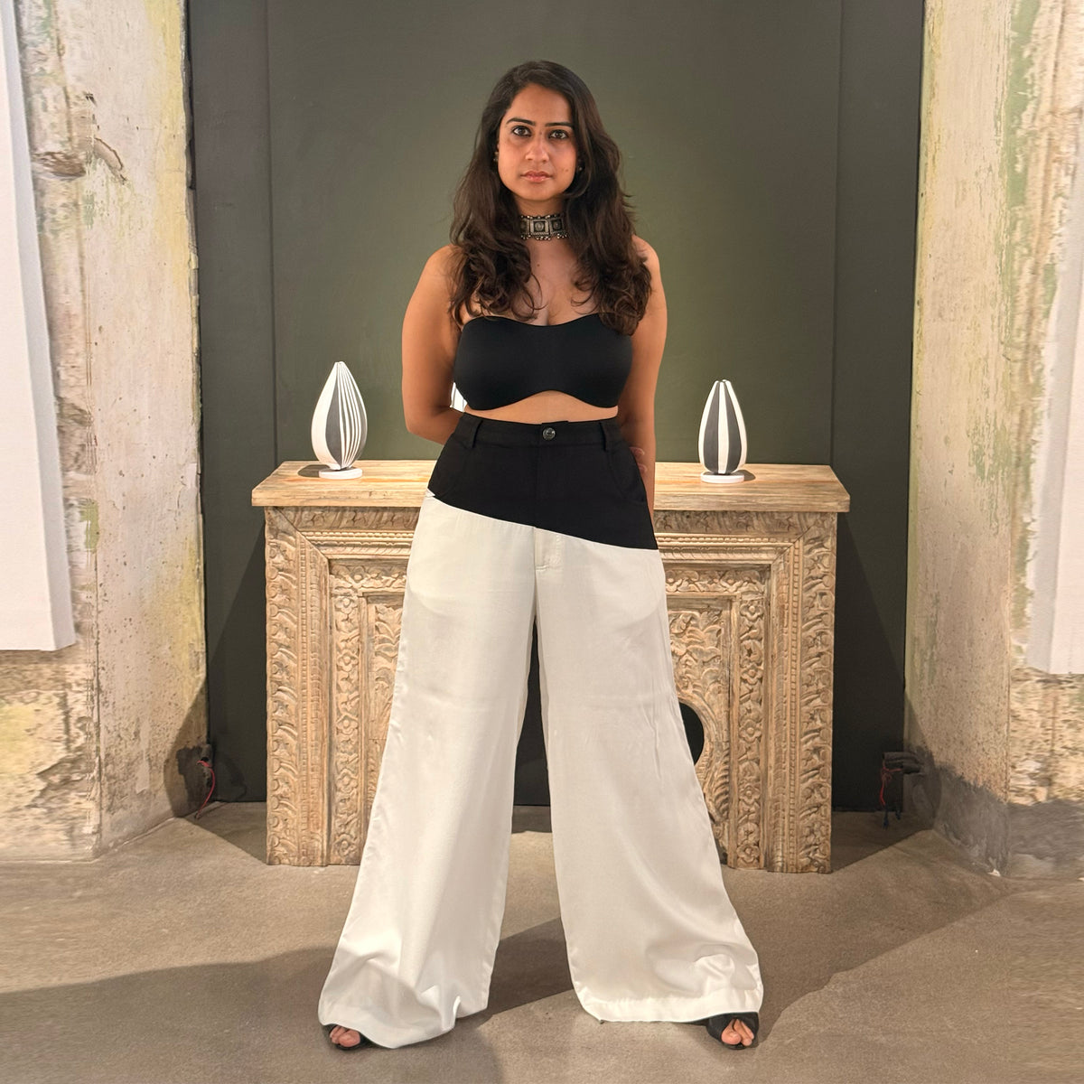Model in white wide-leg Gemini pants and a black crop top standing by a carved stone console table