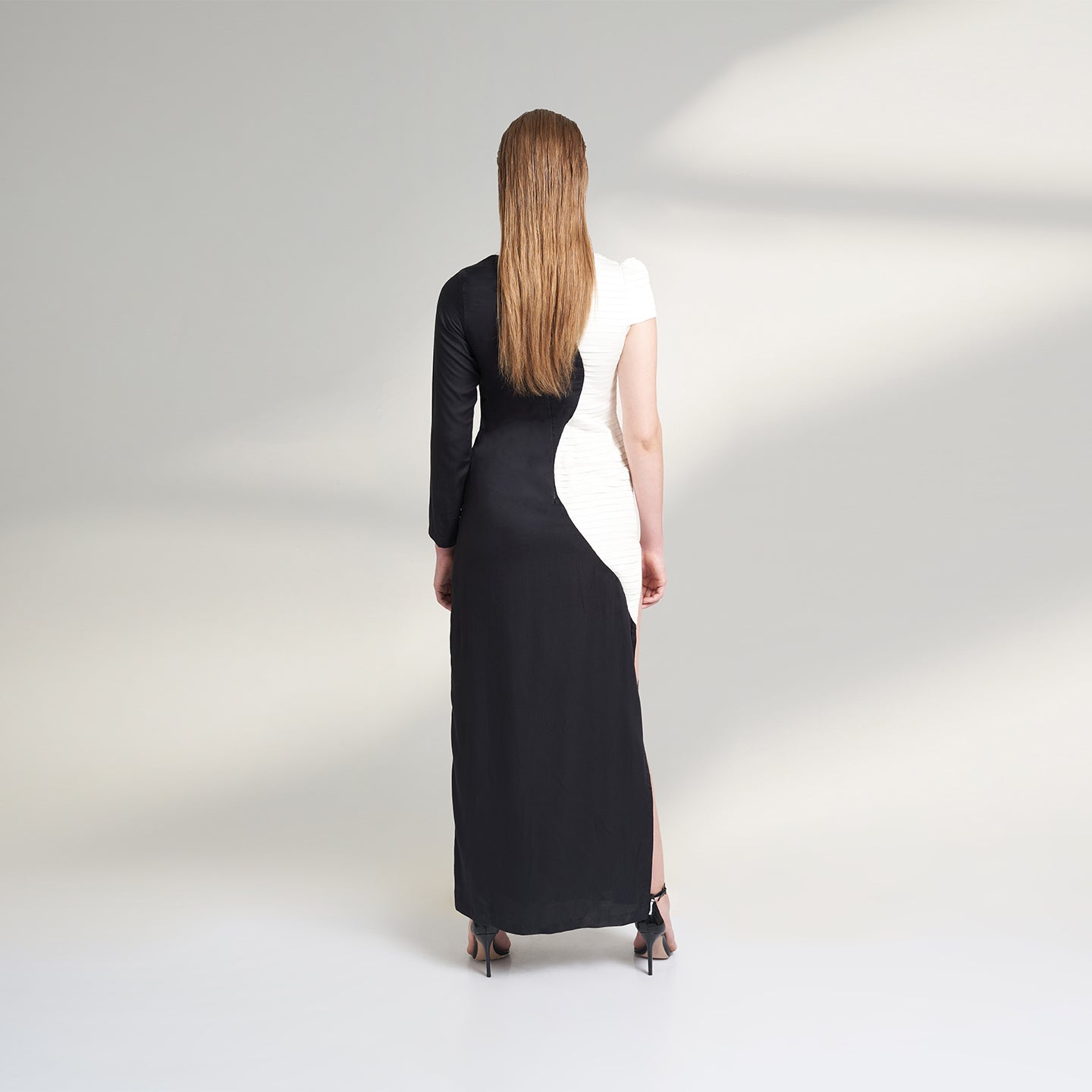 A medium size model wearing a black and white half and half dress made from organic lotus stem fabric. This sustainable vegan dress is a long floor length dress with one side pleated in white and one side plain black with a thigh high side slit striking a back pose.