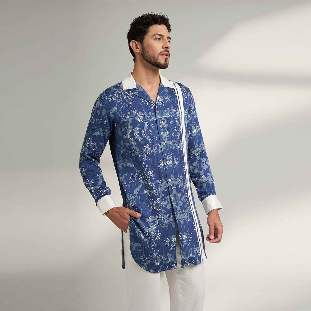 Angled front view of the longline blue floral print shirt with white contrast collar and trim, styled with white trousers